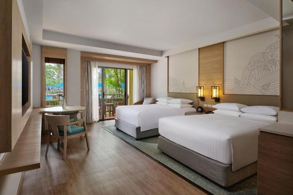 A modern hotel room with two double beds, white linens, a large window with curtains, a balcony with outdoor seating, a small table with chairs, and warm wooden accents.