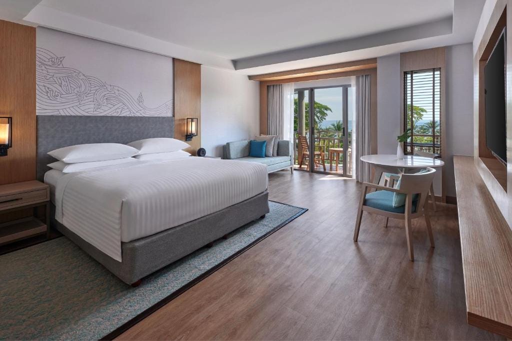 A spacious, modern hotel room with a large bed, sofa, small round table with chairs, wooden floor, wall-mounted TV, and sliding glass doors opening to a balcony with a view of trees and the sea.