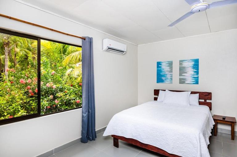 A bright bedroom with a white bed, two abstract blue paintings above the headboard, a wooden nightstand, a ceiling fan, air conditioning, and a window showing lush greenery and pink flowers outside.