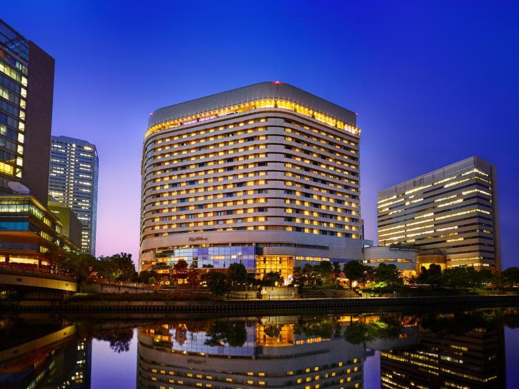 A modern, multi-story hotel with glowing yellow lights stands by a calm river at dusk, flanked by office buildings and reflected in the water under a deep blue sky.