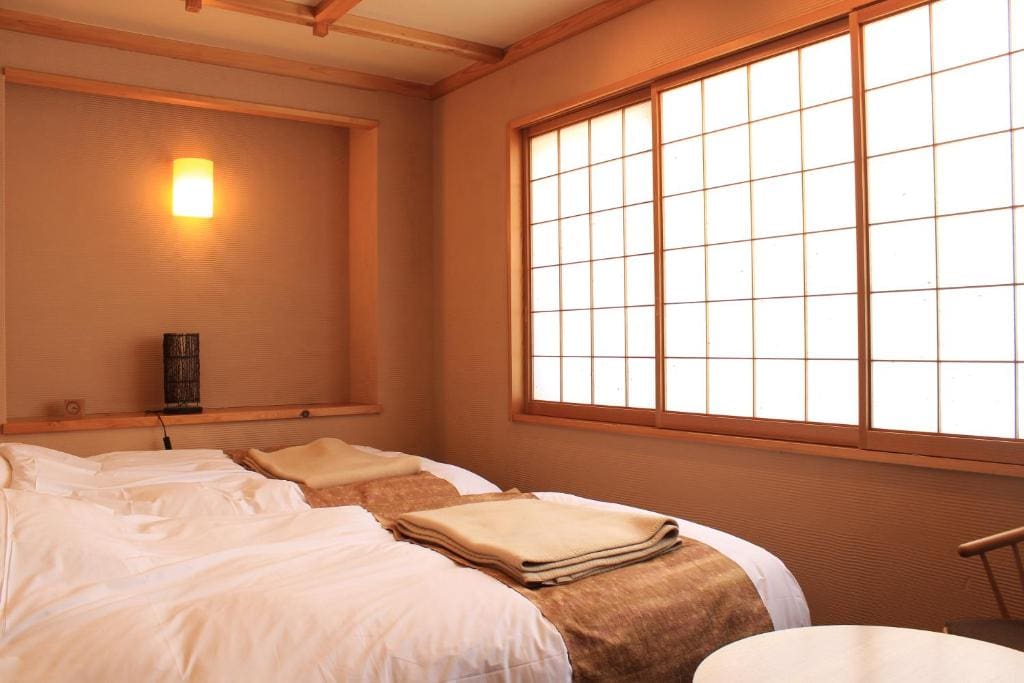 A cozy Japanese-style room with two beds topped with folded blankets, soft lighting, and large shoji paper windows. The decor is minimalist with warm beige tones and natural wood accents.
