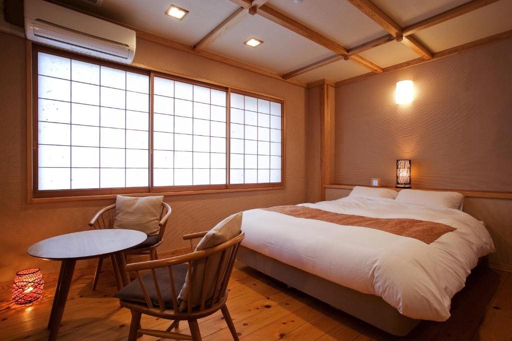 A cozy Japanese-style bedroom with a low bed, white bedding, tatami-inspired floor, wooden furniture, a round table with two chairs, shoji paper windows, and warm ambient lighting.