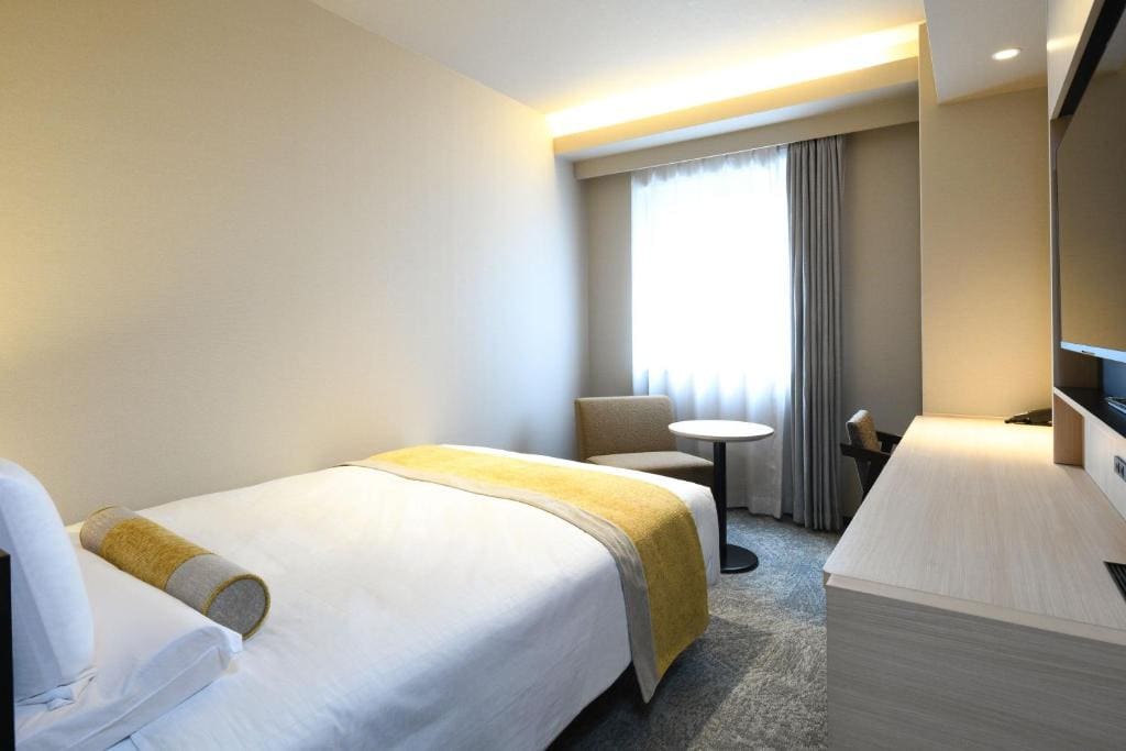 A modern hotel room with a single bed, yellow blanket and bolster, a small round table with a chair by a window with sheer curtains, and a light-colored desk facing a wall-mounted TV.