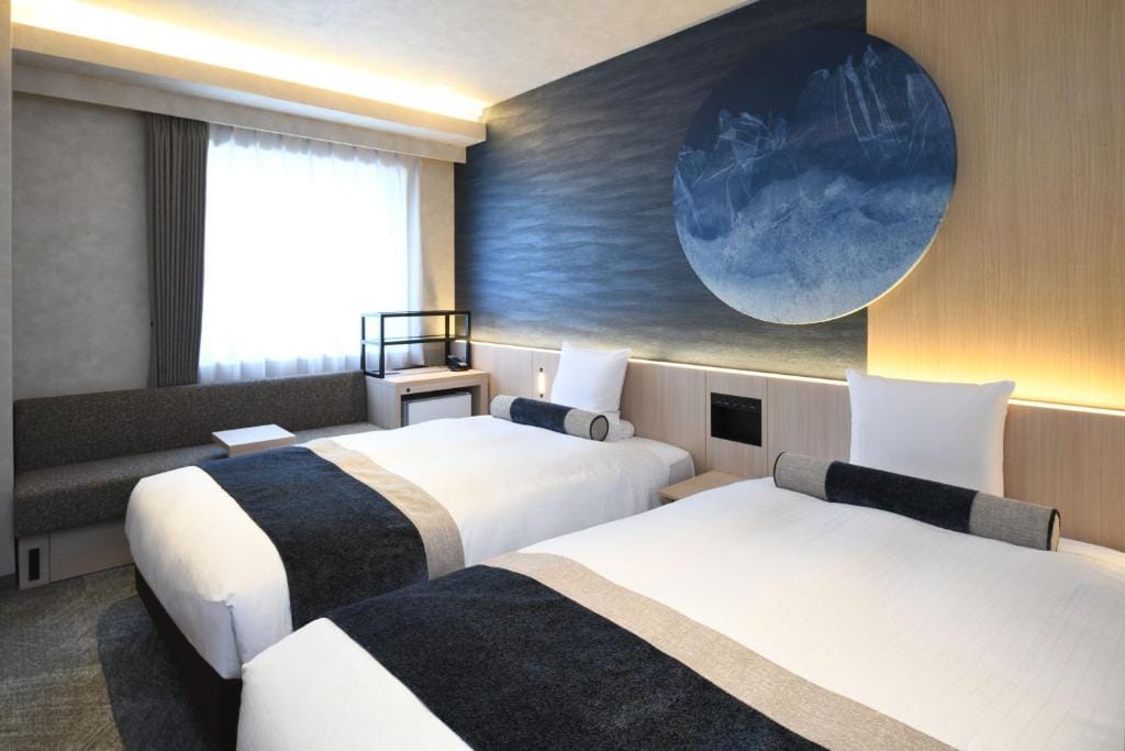 A modern hotel room with two twin beds featuring white linens and dark blue accents, a sofa by the window, and a large circular blue wall art above the headboards. Natural light filters in through a sheer curtain.