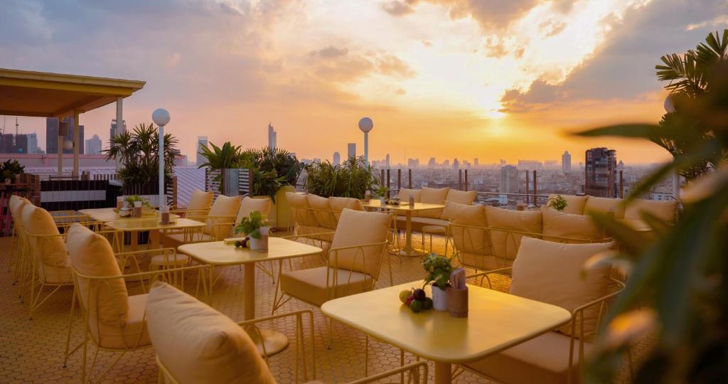 A rooftop restaurant with yellow chairs and tables overlooks a city skyline at sunset. The sky is partly cloudy, and the warm sunlight creates a cozy, inviting atmosphere. Green plants decorate the space.
