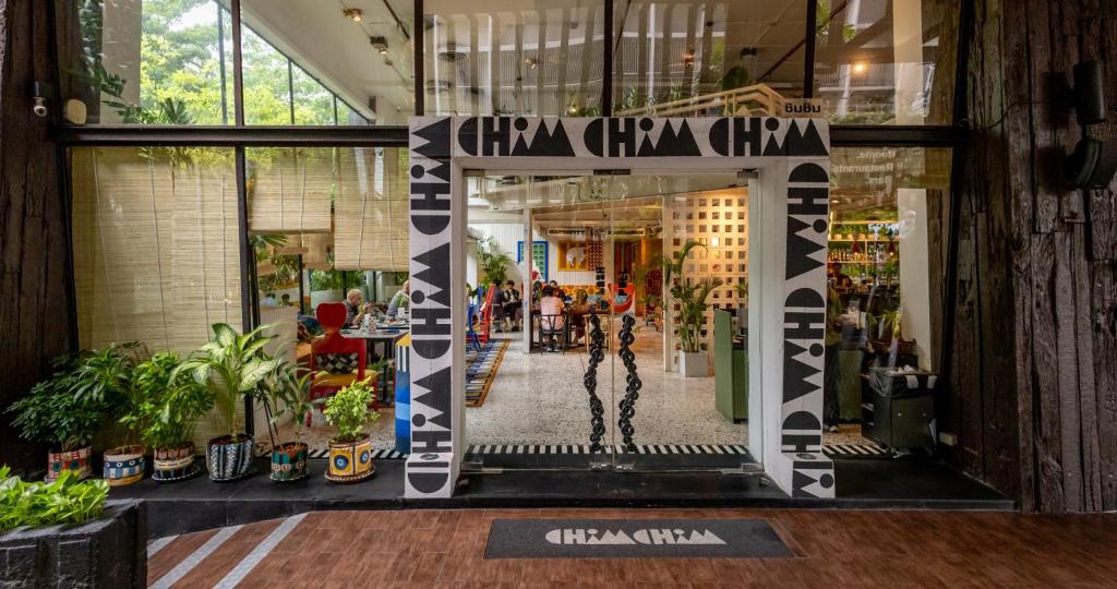 Entrance to a modern café with glass walls, potted plants, and a white doorway decorated with geometric patterns and the word CHÀM. People are visible inside the brightly lit and colorful interior.