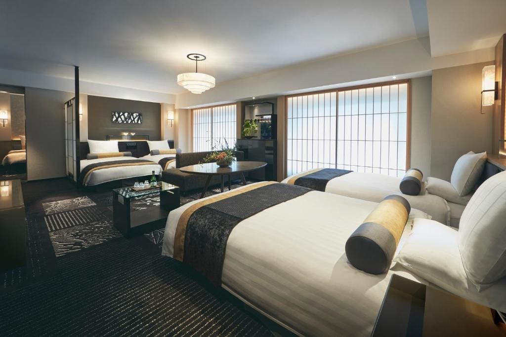 A spacious, modern hotel room with two twin beds, one large bed, stylish decor, a round table with chairs, large windows with blinds, and soft ambient lighting.