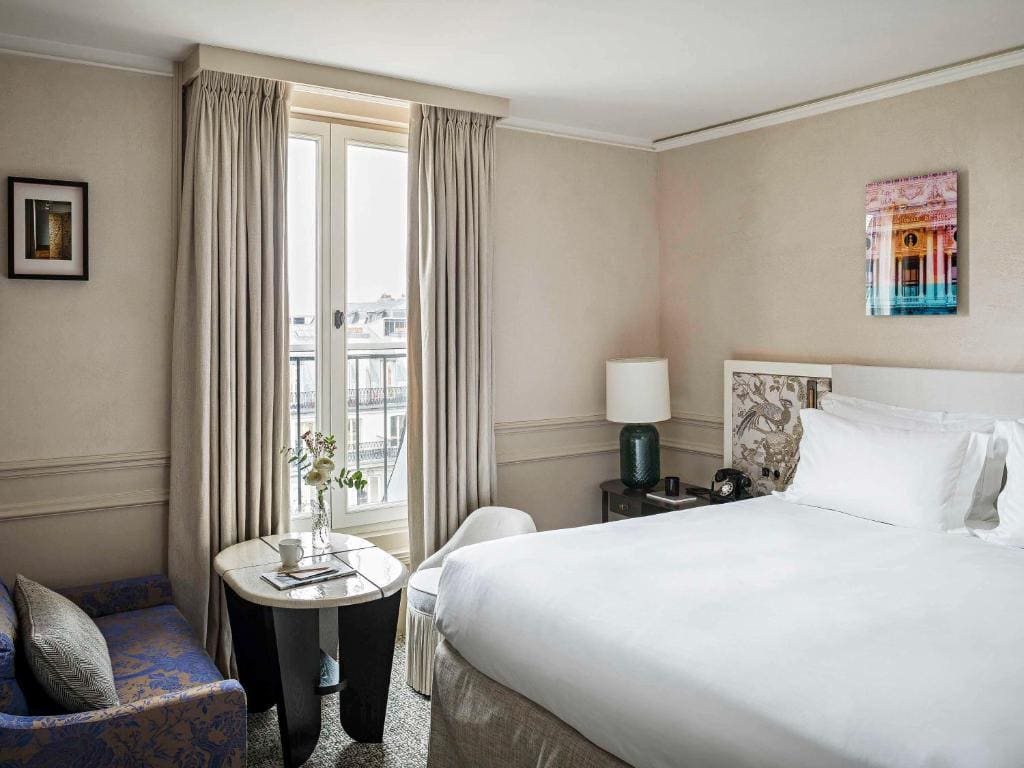 Bright hotel room with a large bed, small table and chair by a window with light curtains, a blue armchair, lamp, and framed artwork on cream-colored walls.