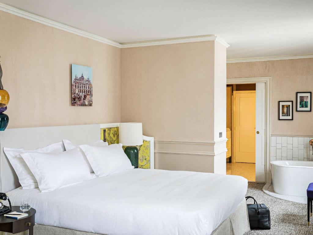 A hotel room with a large bed, white linens, bedside tables, a lamp, framed pictures on the walls, a freestanding bathtub, and an open doorway leading to another room. A black bag sits on an ottoman by the bed.