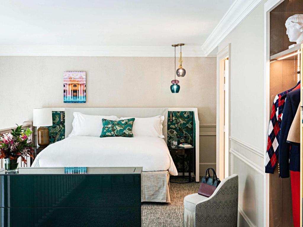 A modern hotel room with a neatly made bed, floral cushions, bedside tables, hanging pendant lights, a flower arrangement, an open closet with clothes, and a small chair near a green dresser.