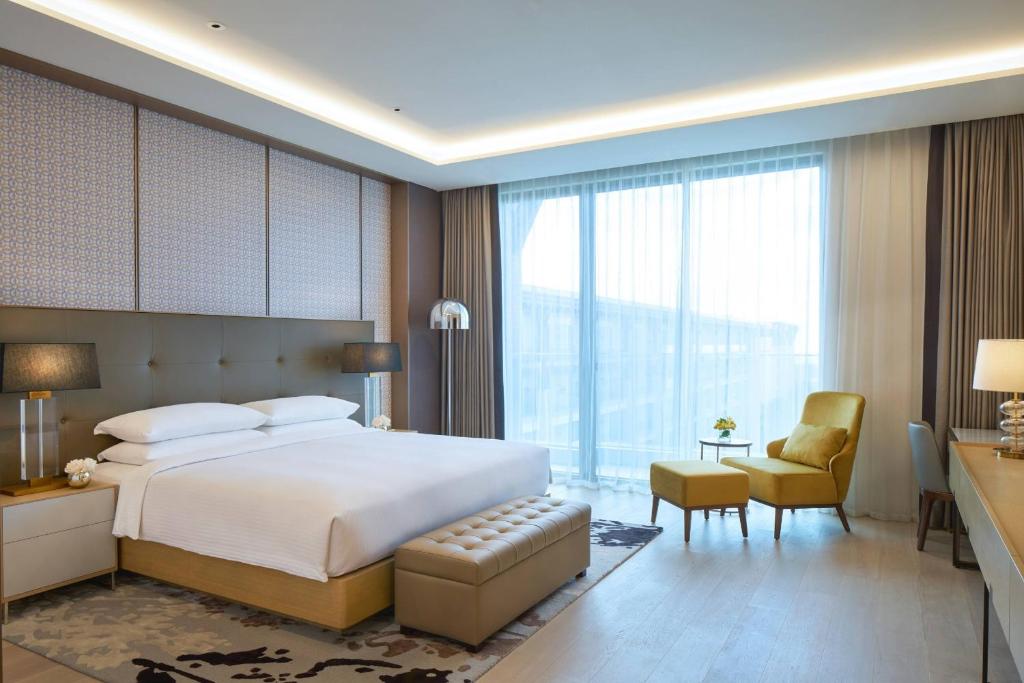 A modern hotel room with a large bed, two bedside tables with lamps, a padded bench, a yellow armchair with an ottoman, a floor lamp, and large windows with sheer curtains allowing natural light in.