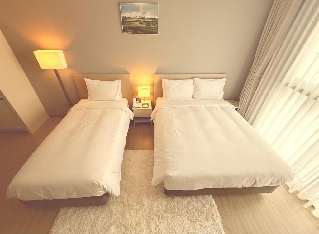 A bright, minimalist bedroom with two neatly made beds, one single and one double, white bedding, a white rug, a nightstand with a lamp and telephone, a floor lamp, and large sheer curtains letting in natural light.
