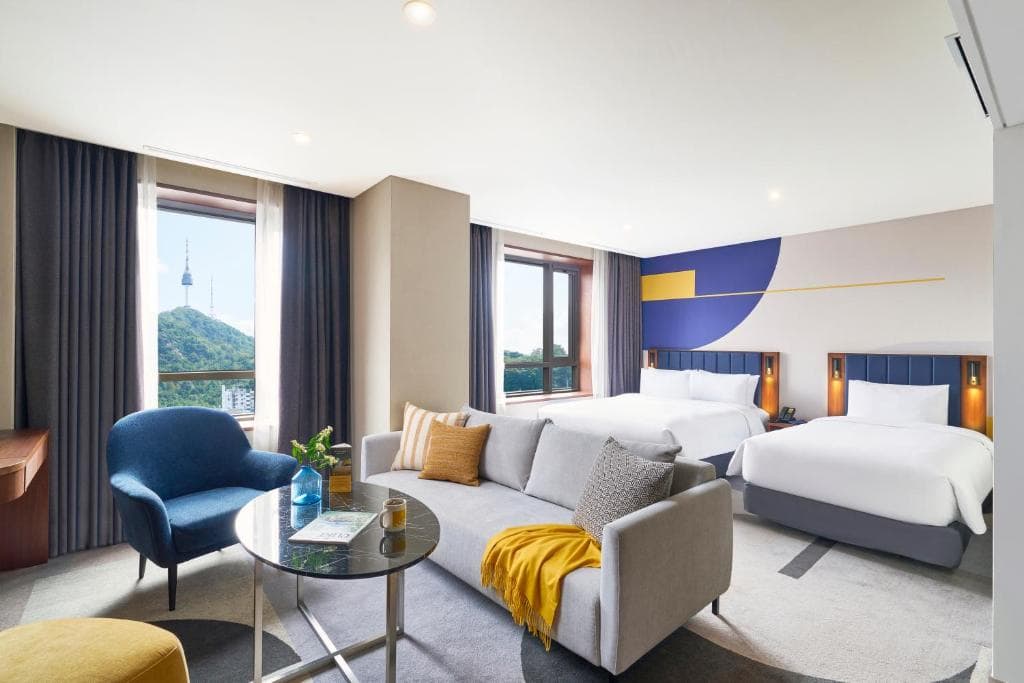 Modern hotel room with two double beds, a gray sofa with yellow accents, a blue armchair, a round glass coffee table, large windows with city and tower views, and stylish decor in blue, gray, and yellow tones.