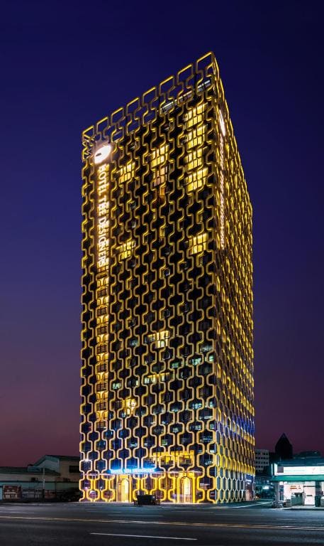 A tall, modern building illuminated at night, featuring a unique yellow geometric grid facade with glowing windows. The sky is dark, and the building stands out brightly among lower surrounding structures.