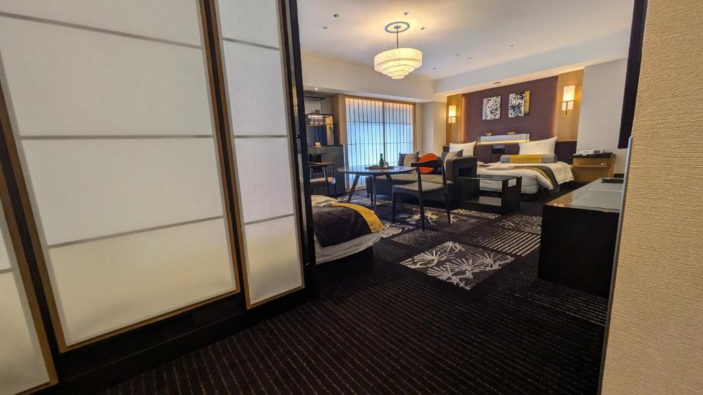 Spacious modern hotel room with two beds, a sitting area, a desk, and contemporary decor. Warm lighting, patterned carpet, and large windows with sheer curtains create a welcoming atmosphere.