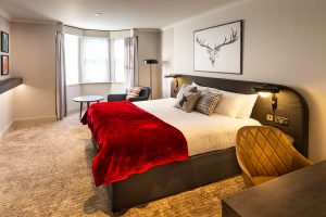 A modern hotel room featuring a double bed with a red blanket, plaid pillows, a desk and chair, a lounge chair by a round table near a bay window, and a framed deer illustration above the bed.