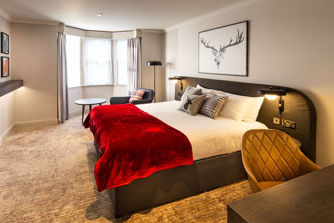A modern hotel room featuring a double bed with a red blanket, plaid pillows, a desk and chair, a lounge chair by a round table near a bay window, and a framed deer illustration above the bed.