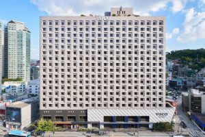 Front view of a modern, high-rise VOCO hotel building with a checkered window pattern, situated in a busy urban area with other buildings, trees, and a clear blue sky in the background.