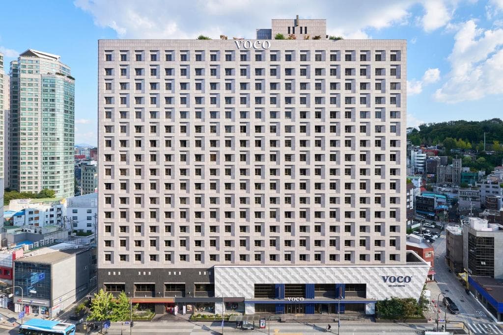 Front view of a modern, high-rise VOCO hotel building with a checkered window pattern, situated in a busy urban area with other buildings, trees, and a clear blue sky in the background.