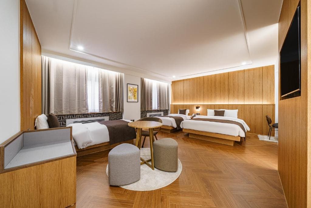 Spacious modern hotel room with two beds, a round table with two stools, wood-panel walls, parquet flooring, a wall-mounted TV, and large windows with curtains letting in natural light.