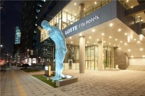 A glowing blue human-shaped sculpture stands in front of the illuminated entrance of LOTTE City Hotels at night, with city lights and tall buildings in the background.