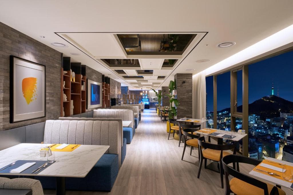 Modern restaurant interior with marble tables, upholstered booths, and yellow-accented chairs. Large windows reveal a nighttime cityscape with a lit-up tower on a hill in the distance. Warm, ambient lighting fills the space.