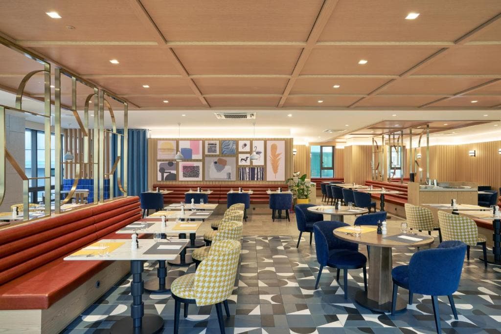 Modern restaurant interior with patterned tile flooring, round and rectangular tables, blue and checkered upholstered chairs, and red banquette seating. Framed artwork decorates the back wall, and the space is well-lit.