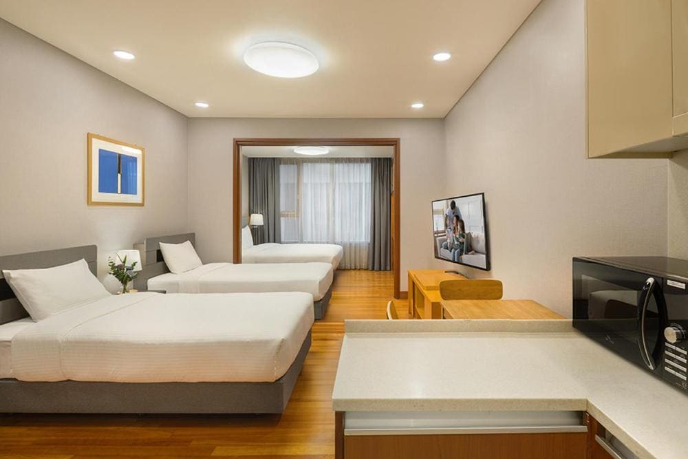 A modern hotel room with two single beds and one double bed, wooden floors, a TV, a microwave, a small table with chairs, and large windows with sheer curtains allowing natural light.