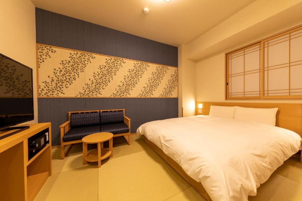 A cozy, modern bedroom with a large bed, a small wooden table with two chairs, a TV on a wooden stand, and a wall decorated with a patterned panel. Warm lighting and a shoji-style window create a peaceful atmosphere.