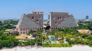 A large, uniquely designed beachfront hotel with two tall, curved towers connected by walkways stands amid lush greenery and pools, with a sandy beach and calm sea in the foreground.