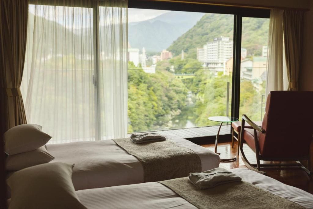 A hotel room with two neatly made twin beds, a red chair, and a small round table, featuring large windows overlooking a lush green landscape and distant mountains.