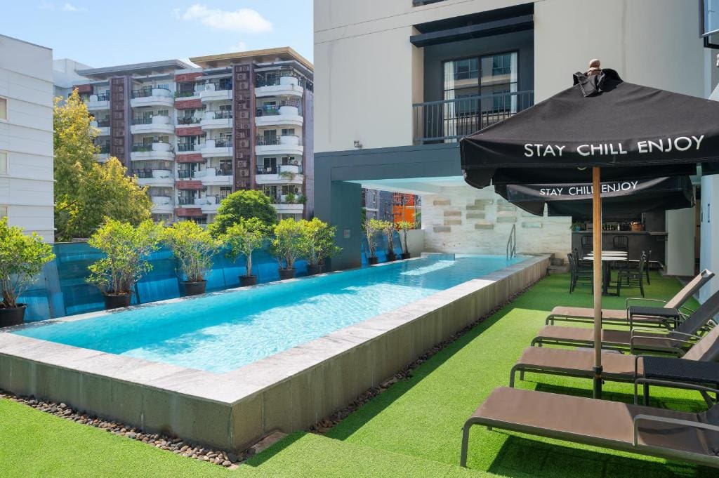 A modern outdoor pool with blue water, surrounded by sun loungers on artificial grass, potted plants along the pool edge, and large black umbrellas that read STAY CHILL ENJOY. Apartment buildings are visible in the background.