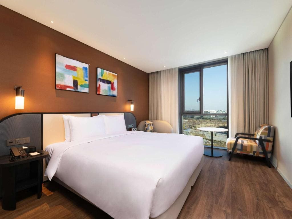 Modern hotel room with a large bed, white bedding, brown accent wall, abstract art, bedside lamps, a window with sheer curtains, two chairs, and a small round table overlooking a cityscape.