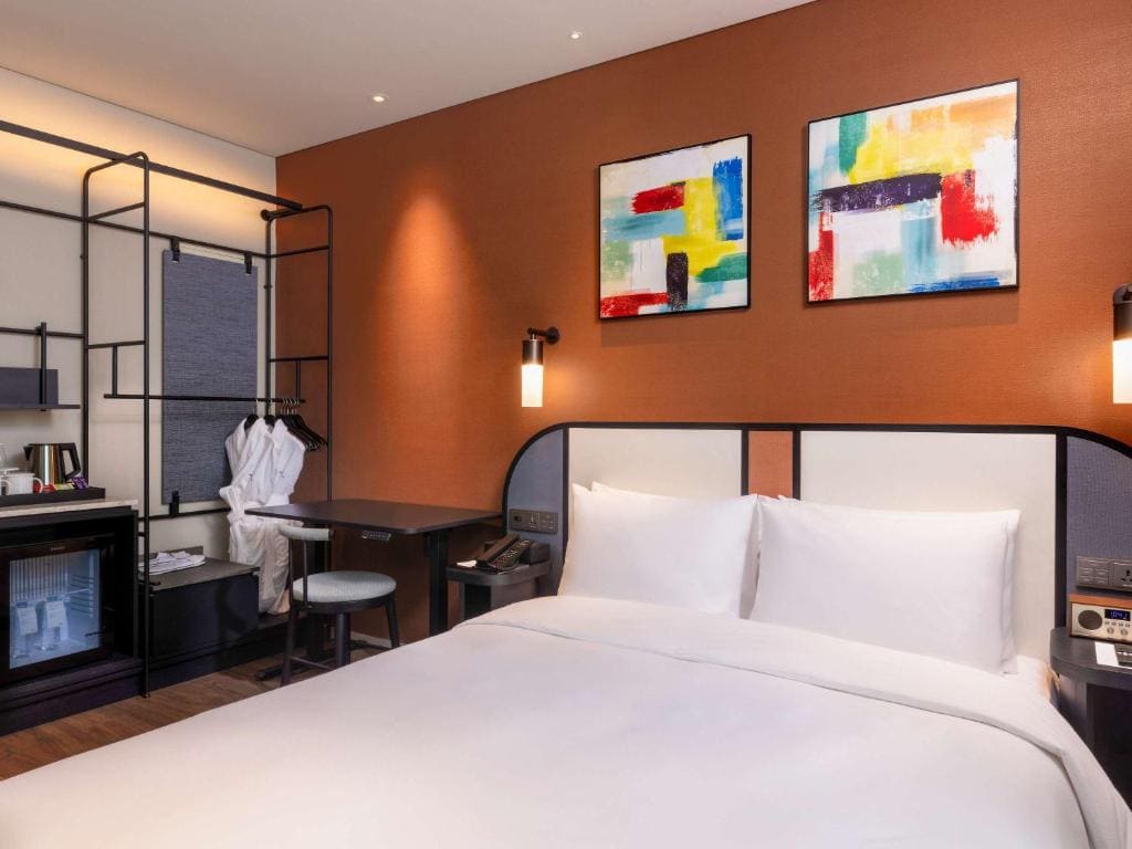 A modern hotel room with a neatly made double bed, two colorful abstract paintings on an orange wall, a small desk with a chair, bathrobes on a rack, and a minibar in the corner.