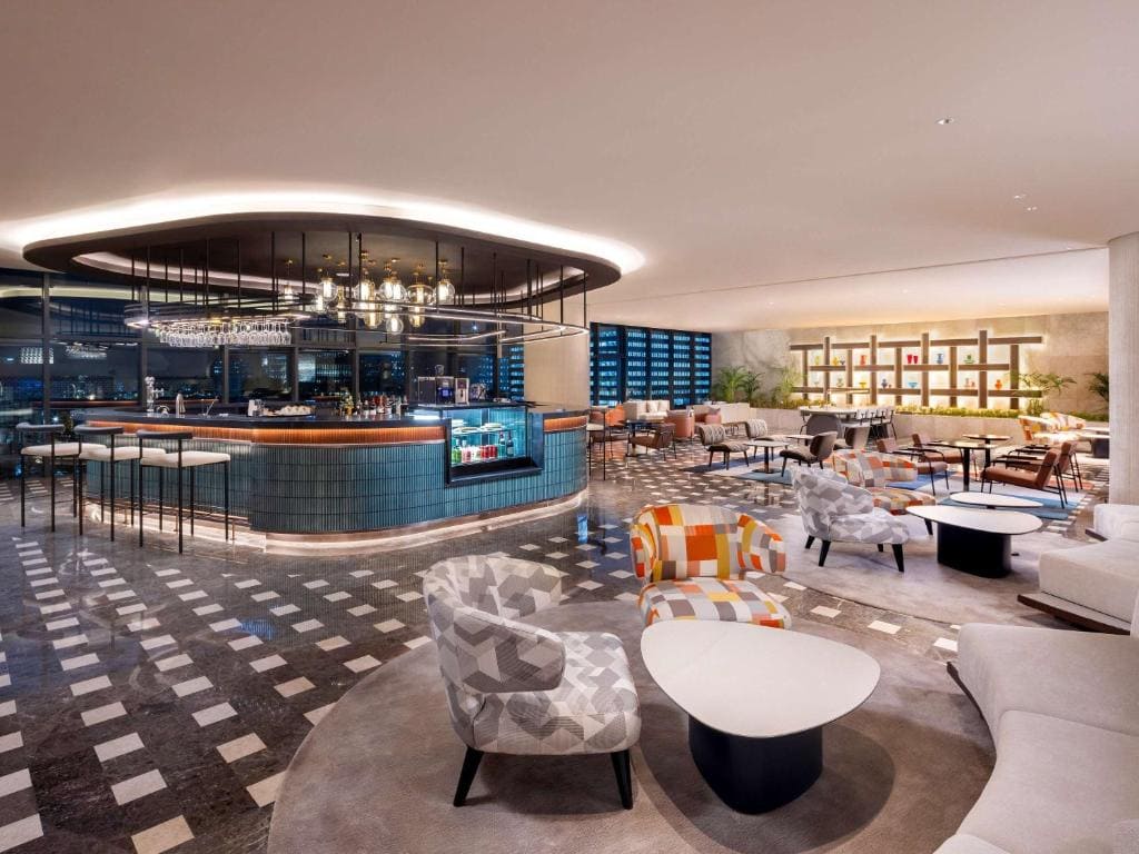 A modern hotel lounge with patterned armchairs, round white tables, tiled floors, and a central circular bar with hanging lights. Shelves and seating areas are in the background, creating a stylish, inviting atmosphere.
