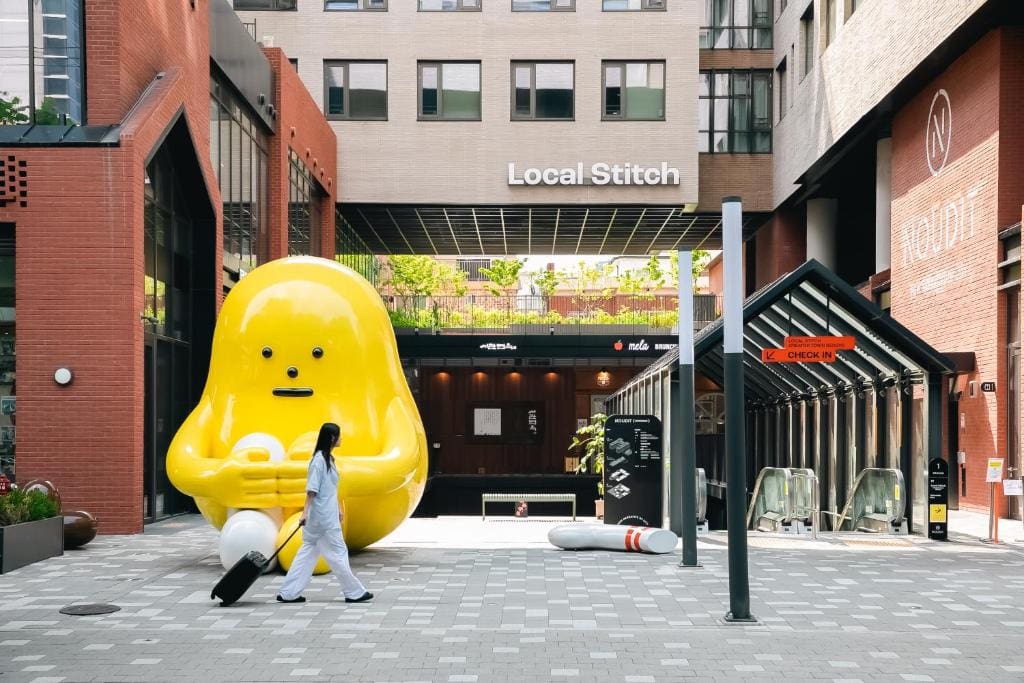 A person in white clothing walks past a large, yellow, cartoon-like statue holding a phone in an urban courtyard, with escalators and the Local Stitch sign in the background.