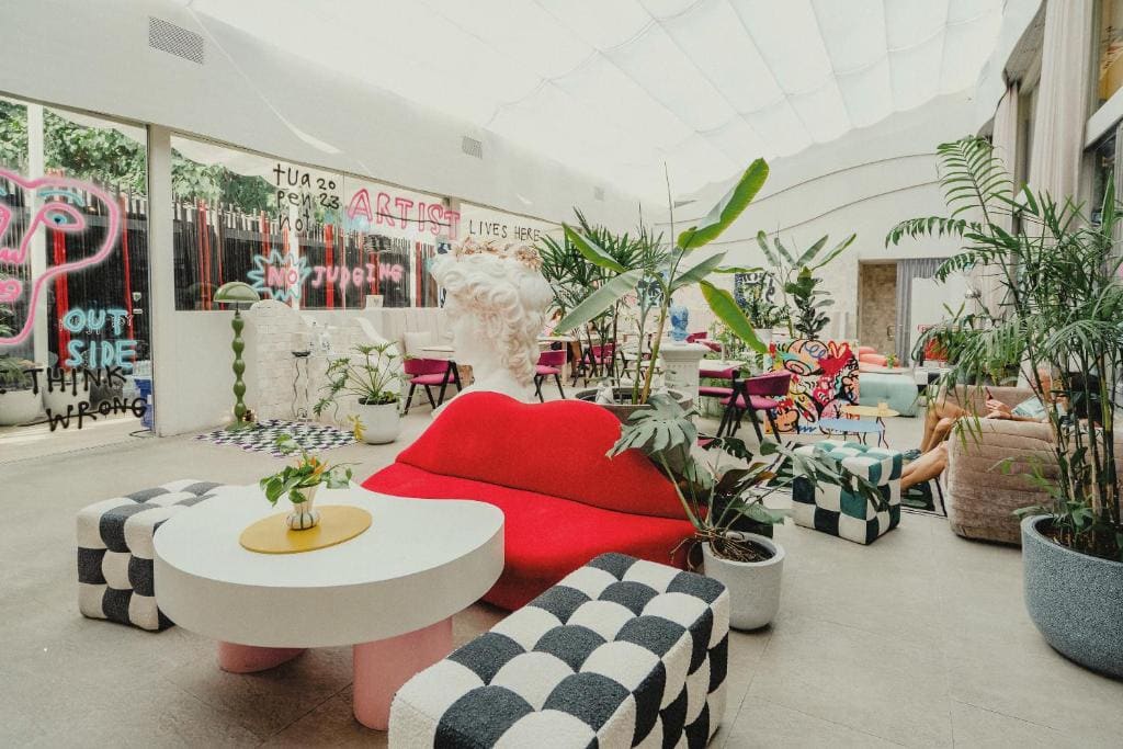 A bright, eclectic lounge features colorful modern furniture, potted plants, a red lip-shaped sofa, checkered stools, playful art, and neon writing on glass walls, creating a vibrant and artistic atmosphere.