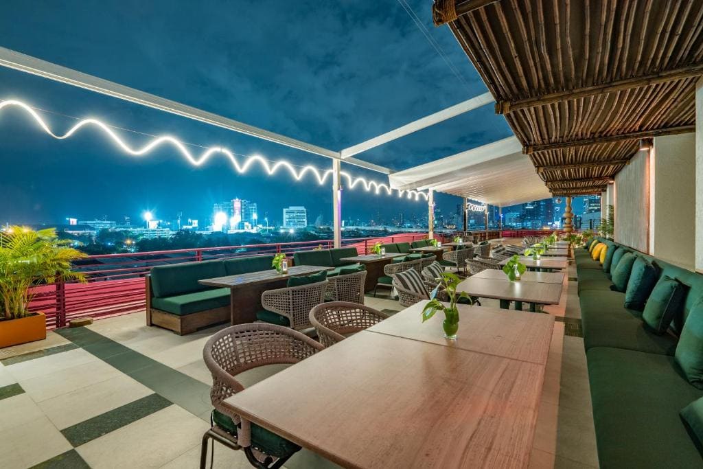 A modern rooftop restaurant with wooden tables, green cushioned seating, potted plants, and a view of a city skyline at night, illuminated by zigzag string lights overhead.