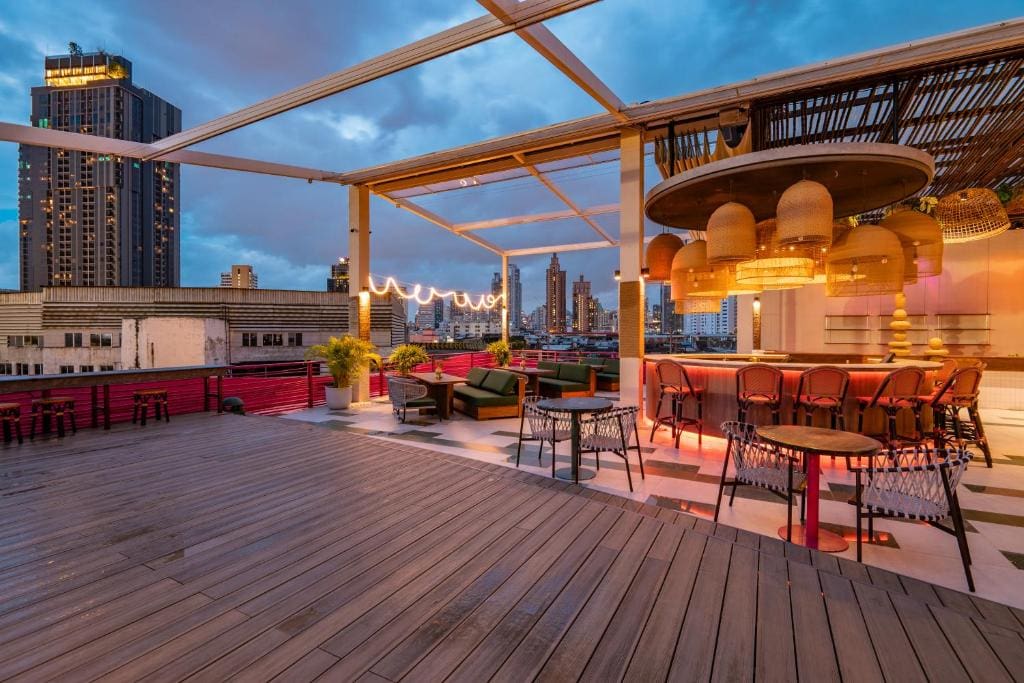 A rooftop bar at dusk with modern furniture, string lights, potted plants, and city buildings in the background under a cloudy sky.