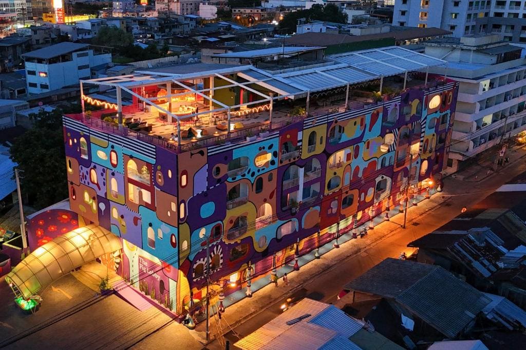 Aerial view of a colorful, multi-story building with abstract murals and bright lights, featuring a rooftop restaurant and surrounded by city streets at dusk.