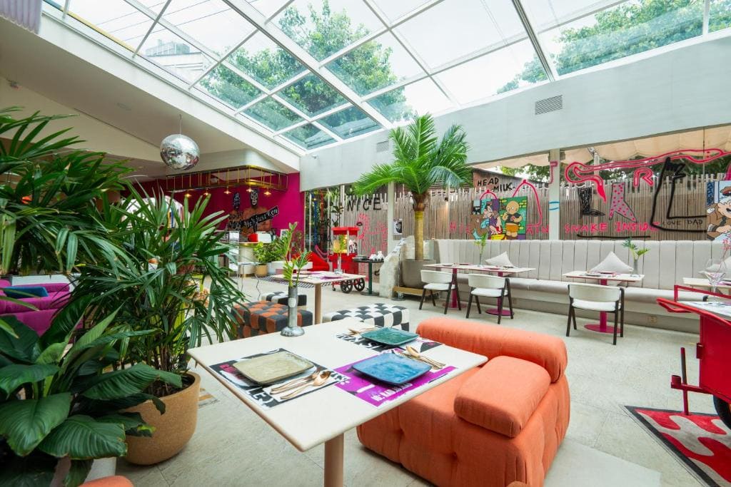 A bright, modern restaurant with large windows, colorful decor, eclectic furniture, indoor plants, and graffiti art on the walls. Tables are set for dining and sunlight streams through the glass ceiling.