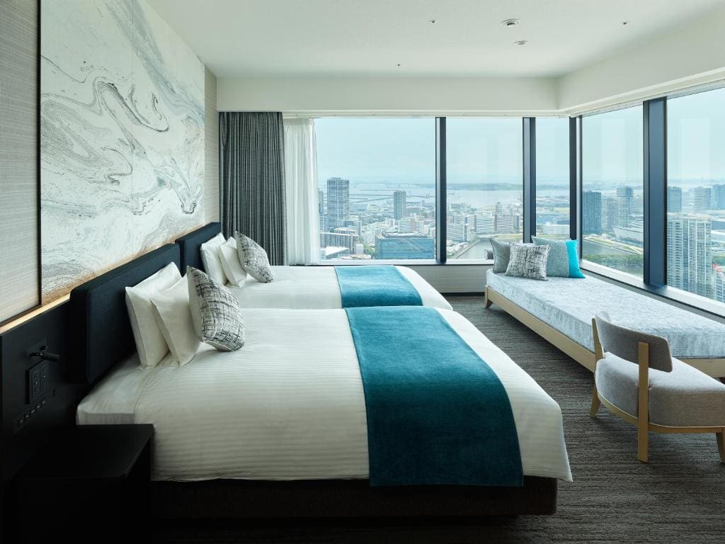 Modern hotel room with two double beds, teal throws, decorative pillows, a marble-patterned accent wall, large windows offering a city and water view, a cushioned window seat, and a wooden chair.