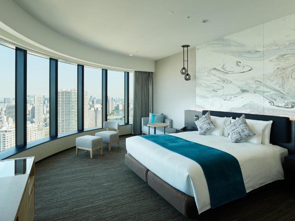 Modern hotel room with a large bed, teal accents, two chairs, and floor-to-ceiling curved windows offering a panoramic city view. Light walls and contemporary decor create a bright, inviting atmosphere.