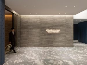 A modern hotel lobby with gray stone walls and floors, featuring a lit sign that reads “Mitsui Garden Hotel Toyosu BAYSIDE CROSS.” A person in a suit walks by, wheeling luggage near an elevator.