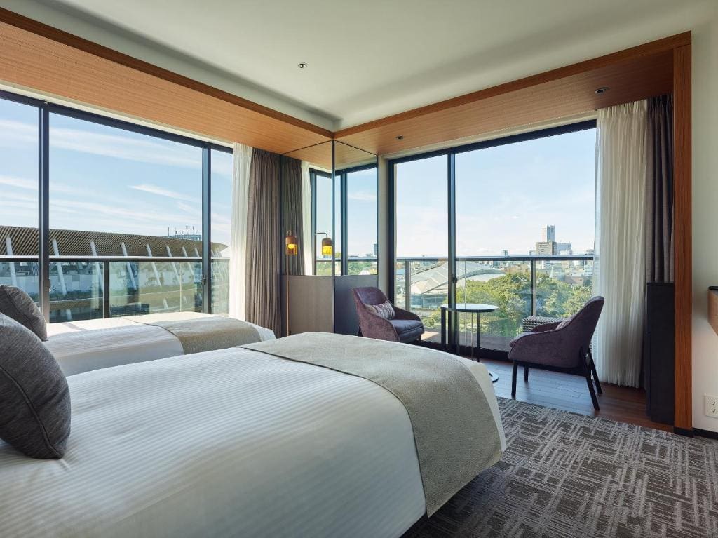 A modern hotel room with two double beds, large windows offering a city view, two chairs and a small round table by the window, and neutral decor with soft natural lighting.