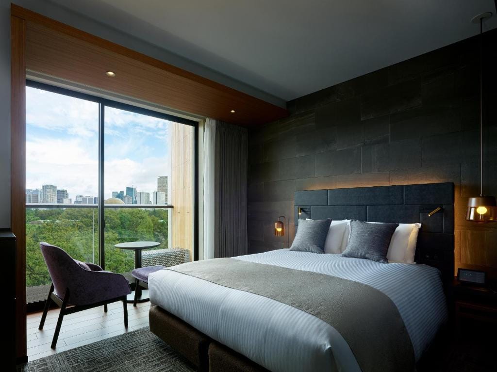 Modern hotel room with a large bed, two bedside lamps, a purple chair, small table, floor-to-ceiling window, and a city skyline view with greenery outside. The room has dark walls and contemporary decor.