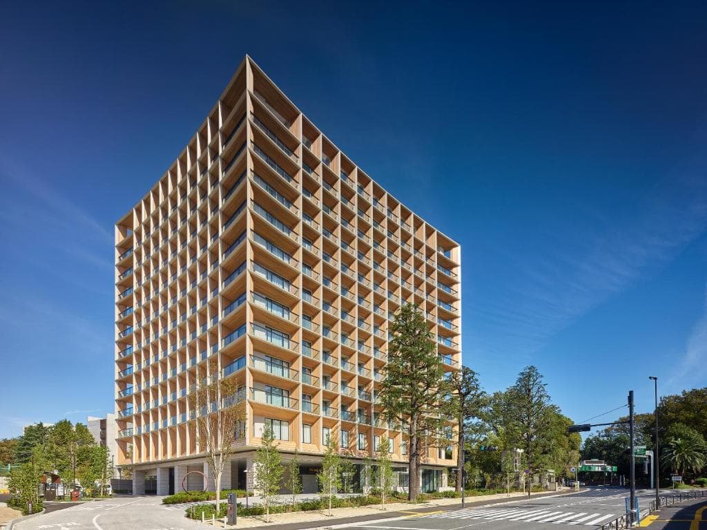 A modern, multi-story building with a unique, angular design and wooden exterior elements stands next to a tree-lined street under a clear blue sky.