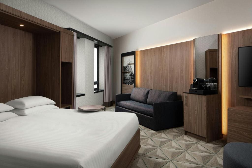 Modern hotel room with a large bed, dark gray sofa, coffee maker on a wooden cabinet, flat-screen TV, and geometric carpet. The room features wood-paneled walls and a window with natural light.