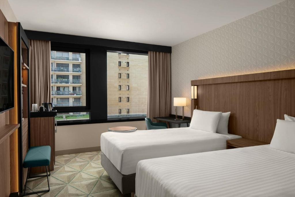 Modern hotel room with two single beds, a wooden headboard, a desk with a chair, a TV, and a window overlooking a building with balconies. Neutral tones and geometric carpet pattern.