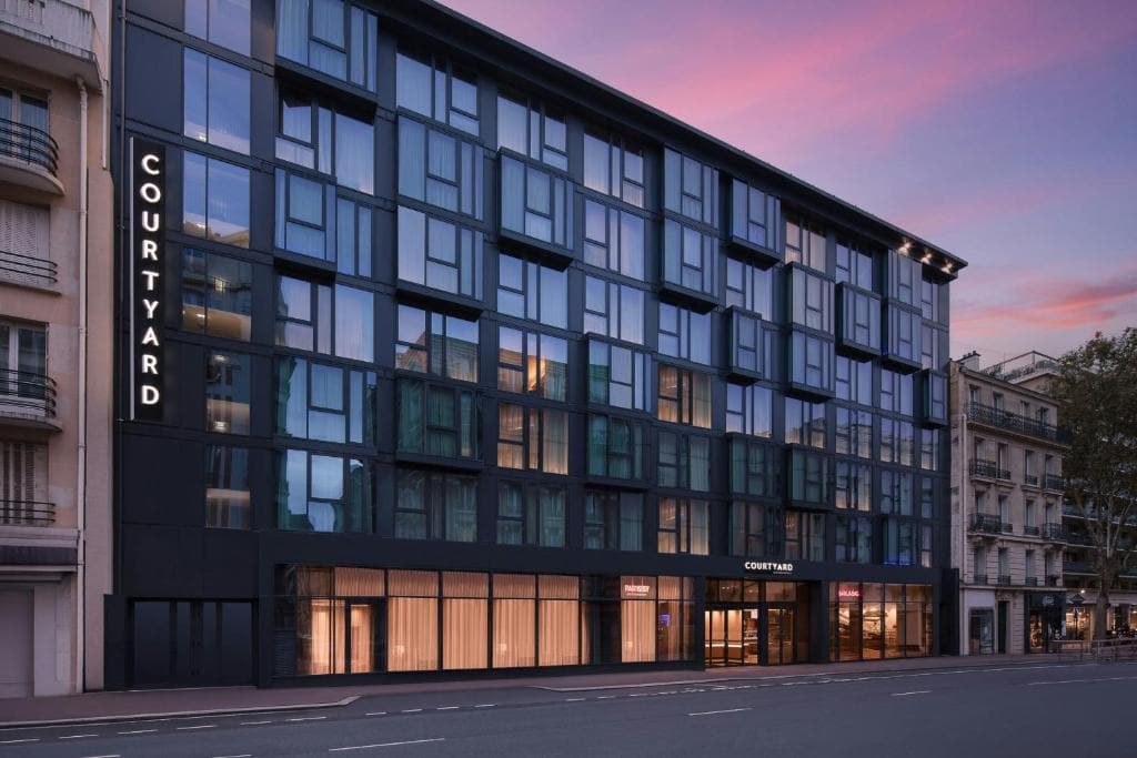 Modern multi-story hotel building with large windows, illuminated signs reading “Courtyard,” and a glass entrance, set along a city street at dusk with a colorful pink and purple sky.